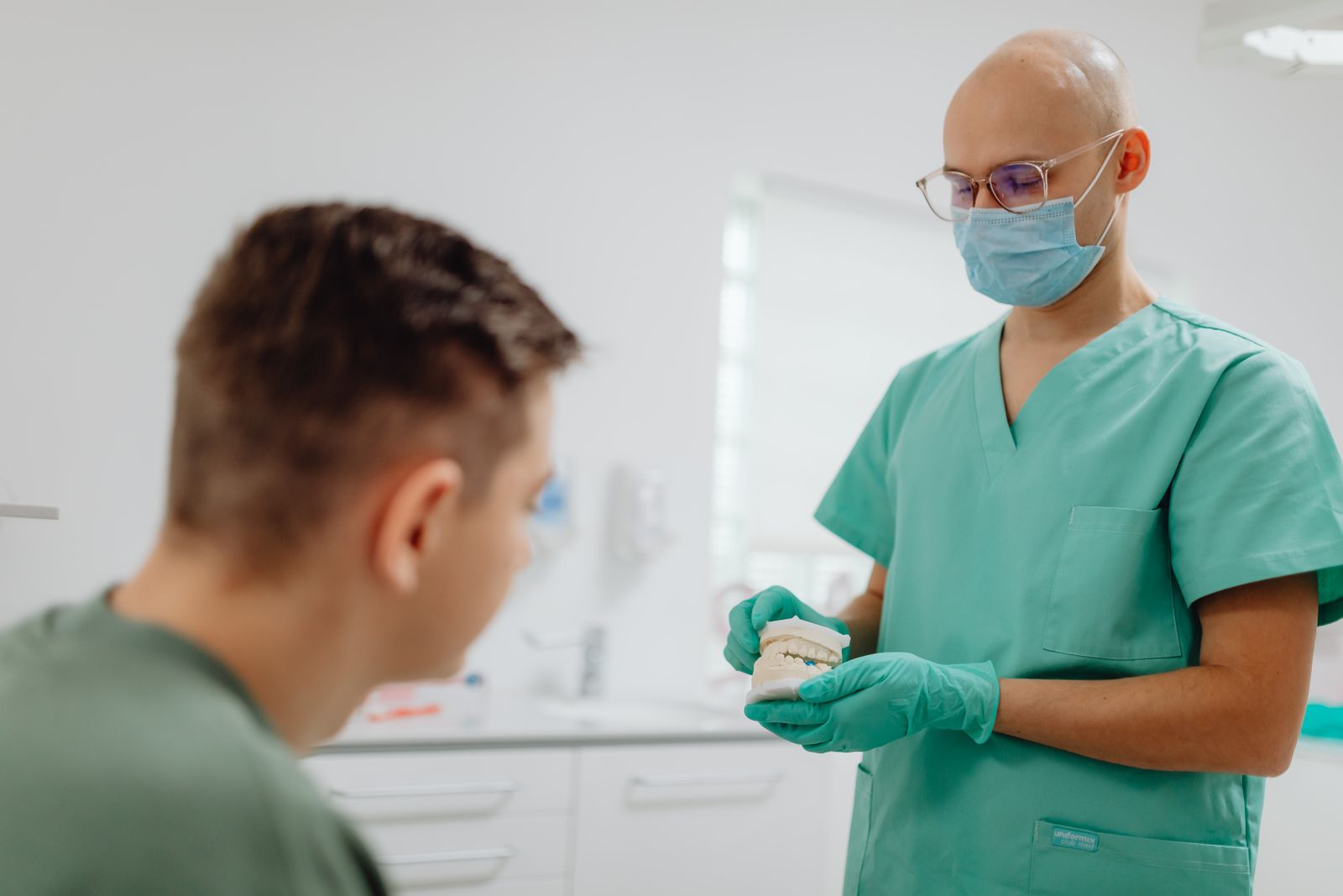 A dentist in scrubs explaining a treatment to a seated patient in a bright clinic