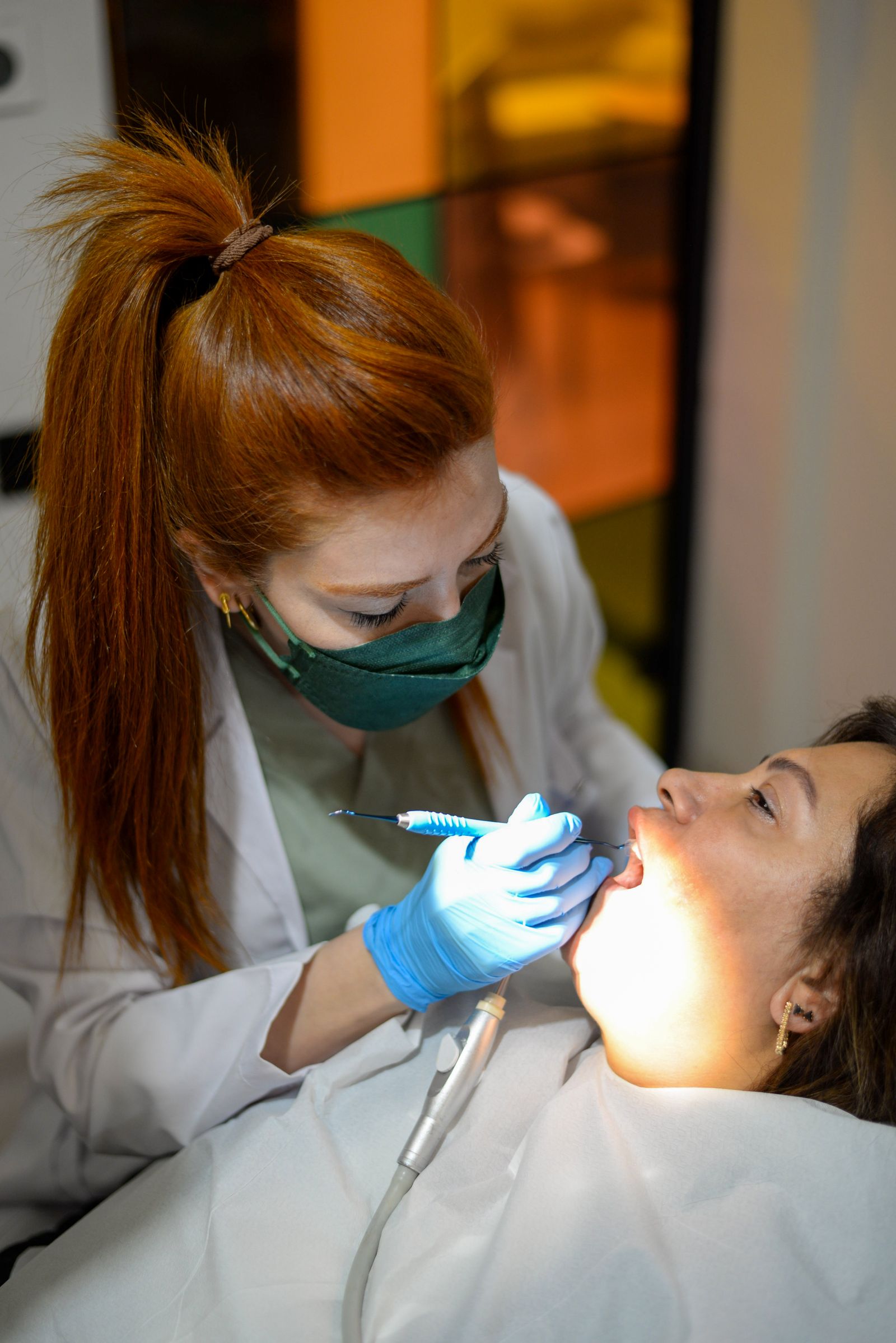 A dentist in a mask and gloves examining a patient's teeth under a bright light